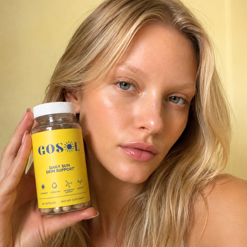 A blonde woman holds a bottle of GOSOL Daily Sun Skin Support dietary supplement in front of her face, set against a soft, warm yellow background.