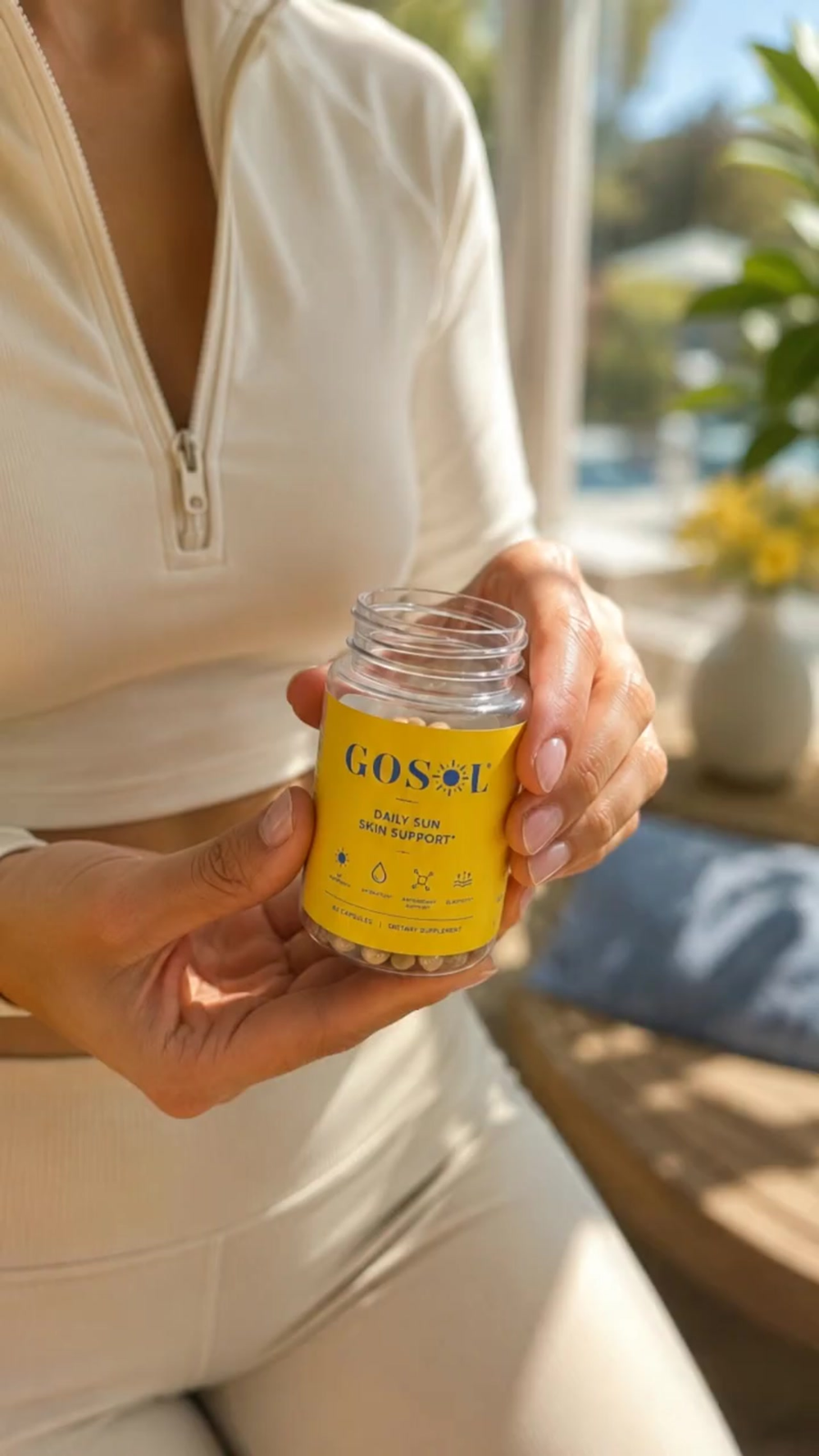 A person in a cream-colored zip-up top holds a small clear jar with a bright yellow label reading COS#L. The setting appears to be a sunlit indoor space with a blurred window, plants, and furniture in the background.