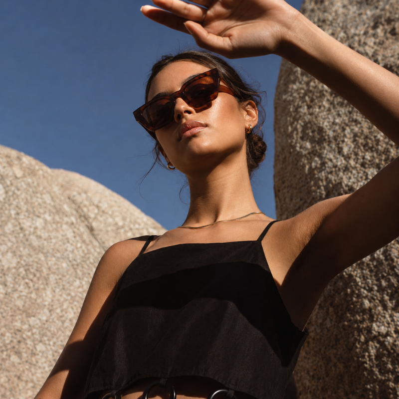 A woman outdoors wearing dark brown cat-eye sunglasses and a black spaghetti-strap top, posing in front of large rock formations under a clear blue sky.
