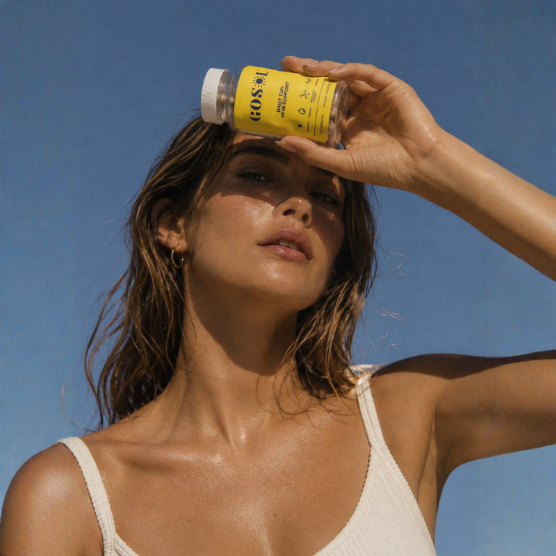 A woman outdoors under a clear blue sky holds a yellow GoSol bottle to her forehead to shield her eyes from the sun. She is wearing a white ribbed tank top.